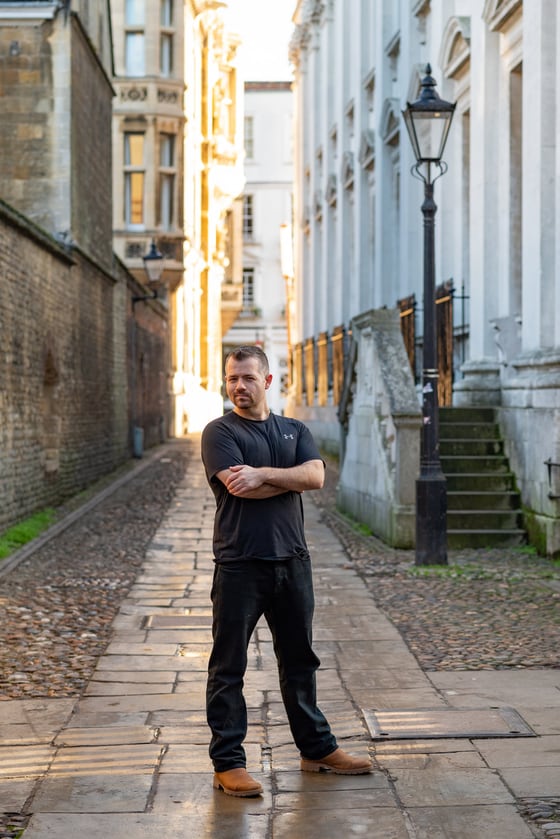 Nick Jacob standing with folded arms in a narrow cobbled lane in Cambridge, between old stone buildings, with an iron street lamp to one side and low sun at the far end of the lane.