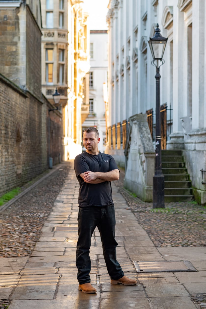 Nick Jacob standing with folded arms in a narrow cobbled lane in Cambridge, between old stone buildings, with an iron street lamp to one side and low sun at the far end of the lane.